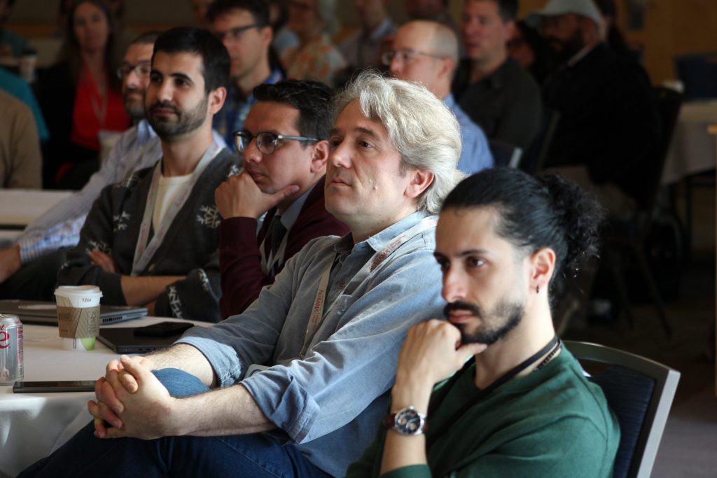 A close-up photo of four conference attendees in different postures, engaged by an off-camera presenter.