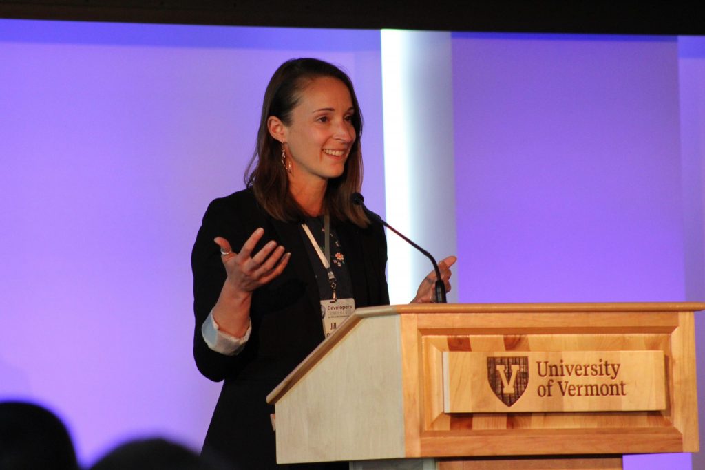 A woman speaking at a wooden podium with the University of Vermont logo, addressing an off-camera audience.