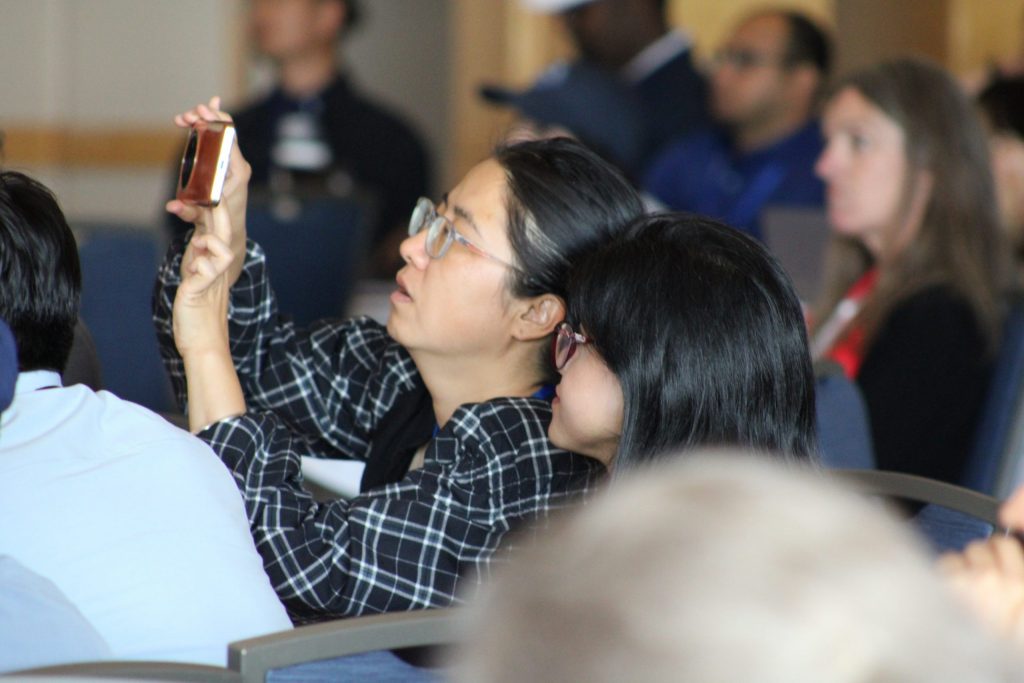 An audience member using their phone to take a photo of a presentation, with other attendees blurred in the background.