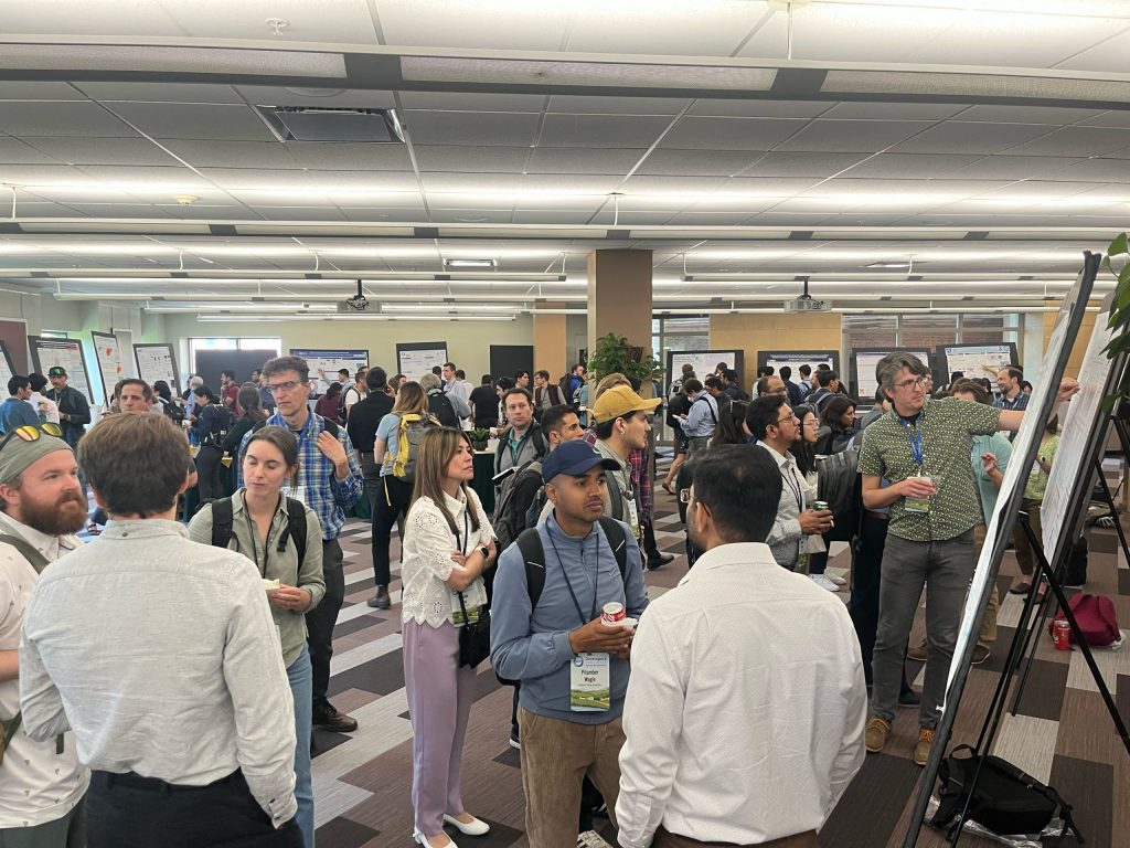 Many conference attendees standing during a poster session, with posters and people in the background, several groups engaged in discussion and people gesturing to posters.