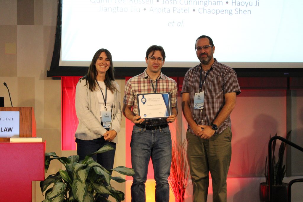 Photograph of three people standing on a stage during an award ceremony, with one person holding a certificate.