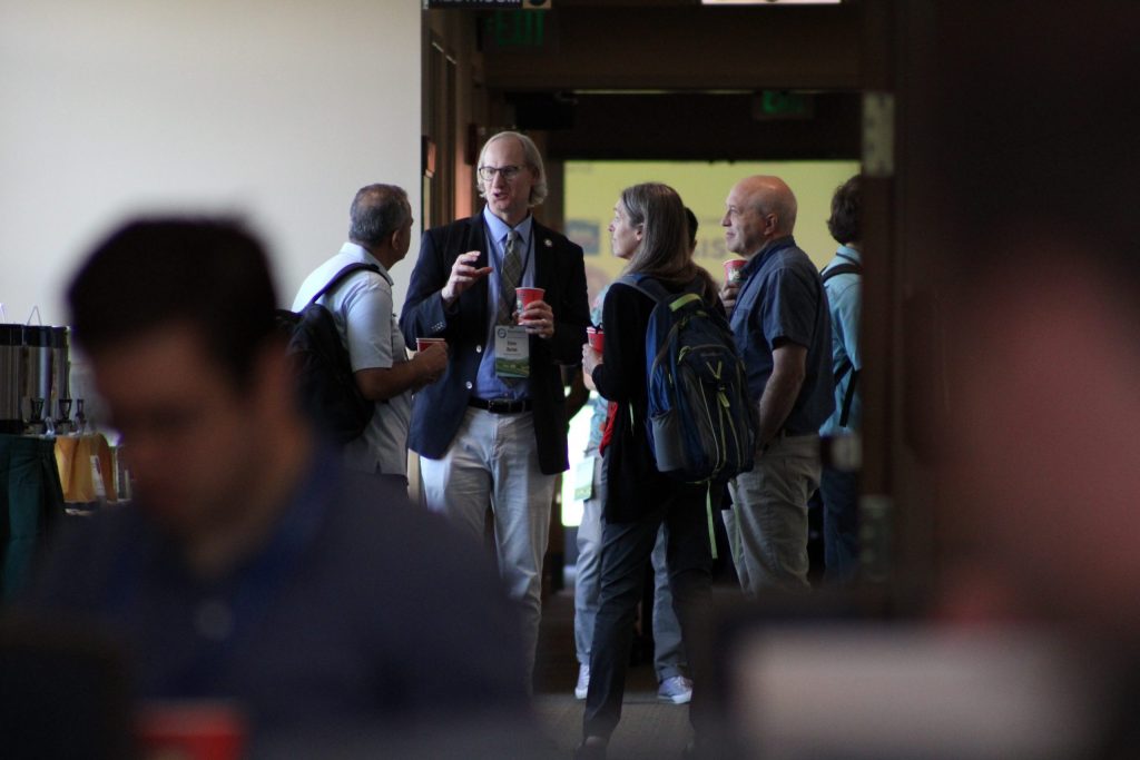 A candid photo of four people holding coffees and engaged in discussion in the midground, with people blurred in the foreground.