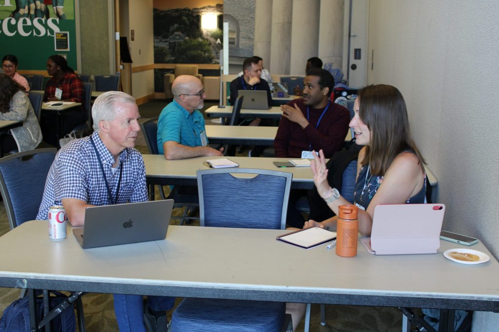 Pairs of conference attendees sitting at tables with laptops open and notebooks out participating in conversations.