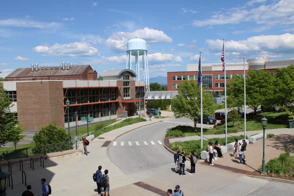 A wide shot that shows the scope of the conference complex outside, capturing a water tower in the background, buildings, flags and landscaping in the midground, and people standing around in the foreground.