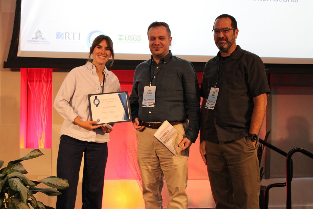 Photograph of three people standing on a stage during an award presentation, with one person holding a framed certificate.