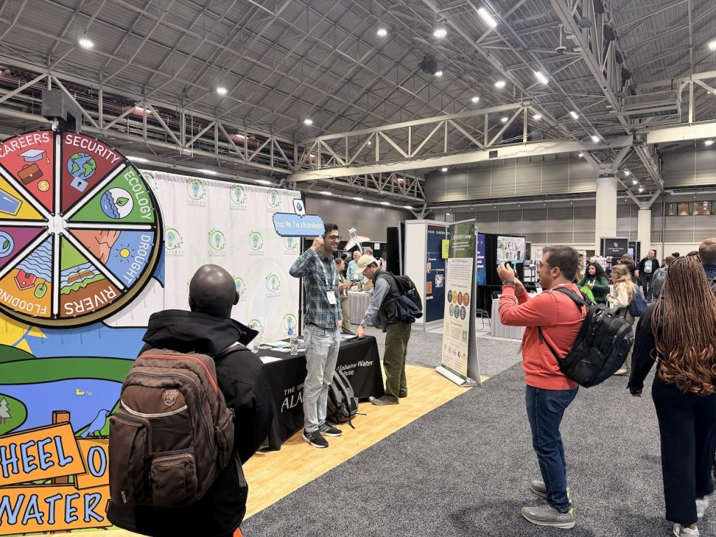 An attendee holds a “Trust me, I’m a hydrologist” sign beside the Wheel of Water as others take photos at the CIROH booth