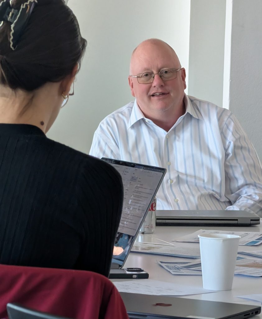 A man speaks during a small meeting while another participant takes notes on a laptop.