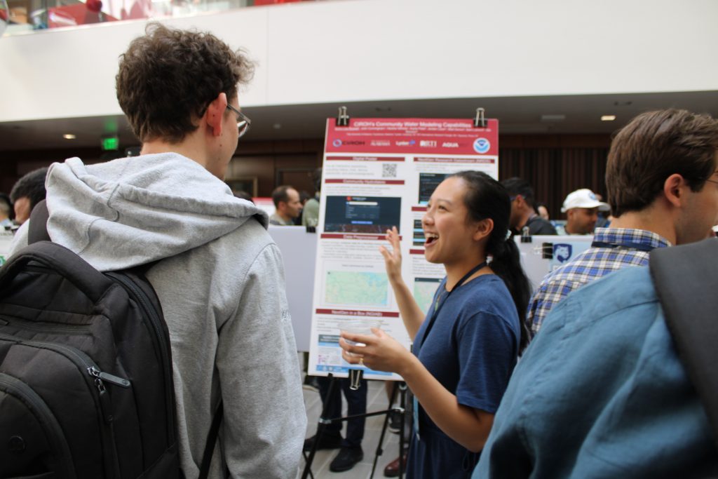 Photograph of a science fair or academic conference showing a presenter explaining a research poster to an attendee.
