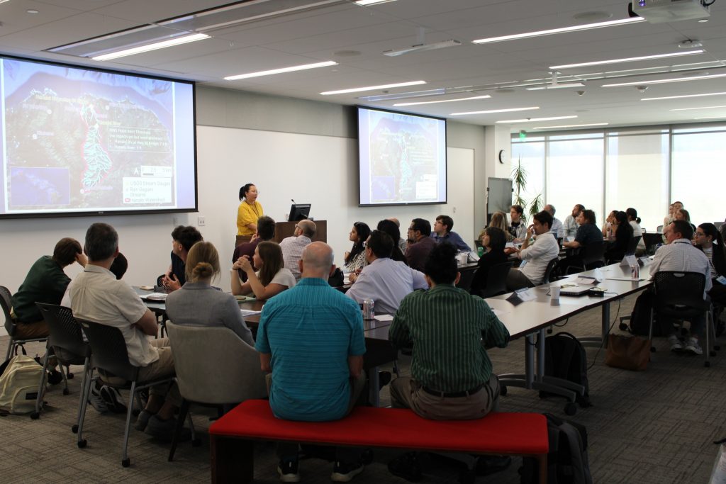 Photograph of a conference room with attendees seated around tables, listening to a presenter standing near two projection screens displaying maps and data.