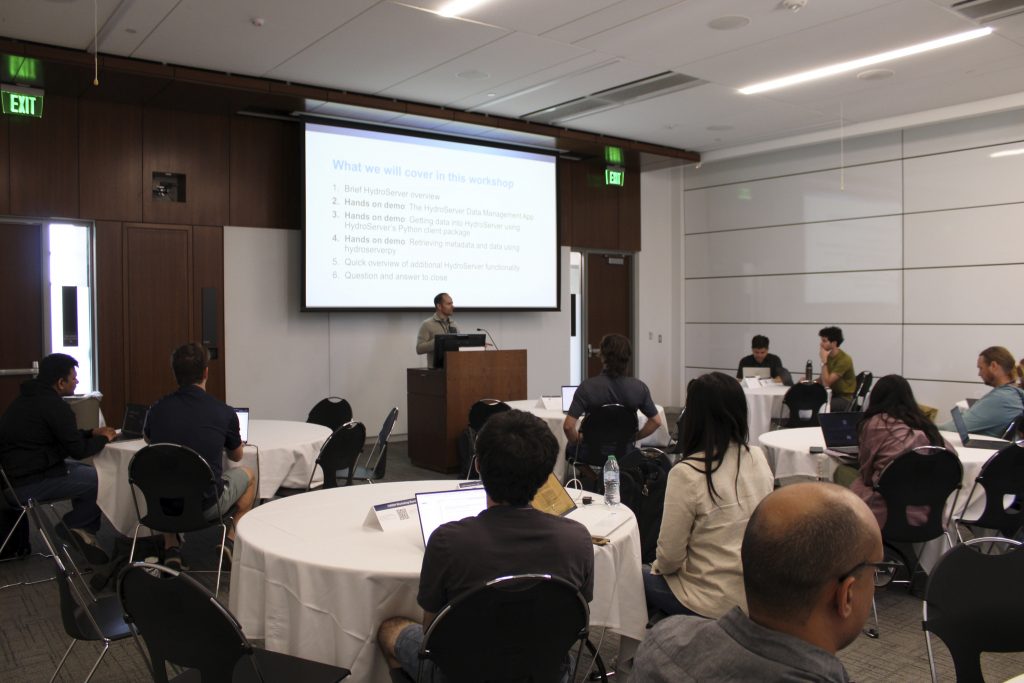 Photograph of a workshop setting with participants seated at round tables facing a presenter standing at a podium in front of a projection screen.
