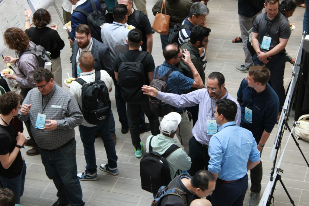 Photograph showing a group of people gathered indoors with many wearing name badges and carrying backpacks