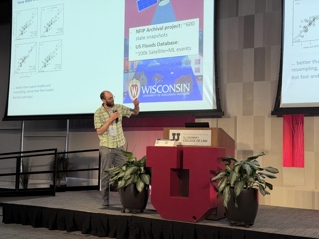 Photograph of a speaker presenting at a conference in a lecture hall, standing next to a podium with "S.J. Quinney College of Law" and a large red "U" emblem.