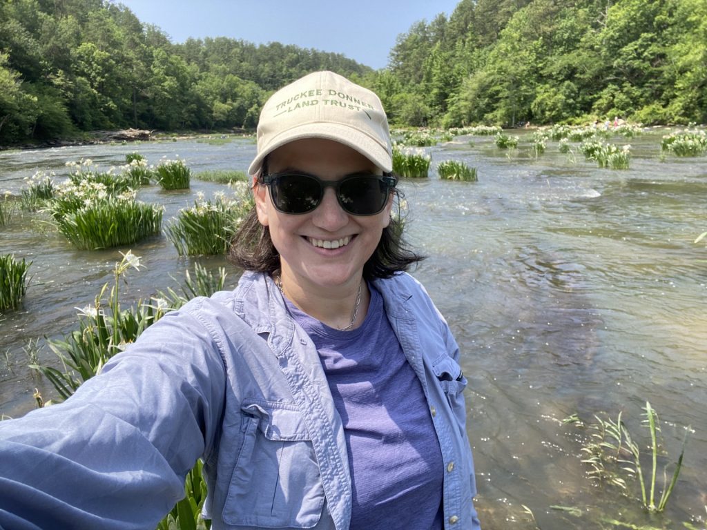 Brauman takes a photo of herself standing at the Cahaba River National Wildlife Refuge, with the river and blooming Cahaba lilies visible behind her. She is smiling towards the camera and dressed in a blue jacket with sunglasses on.