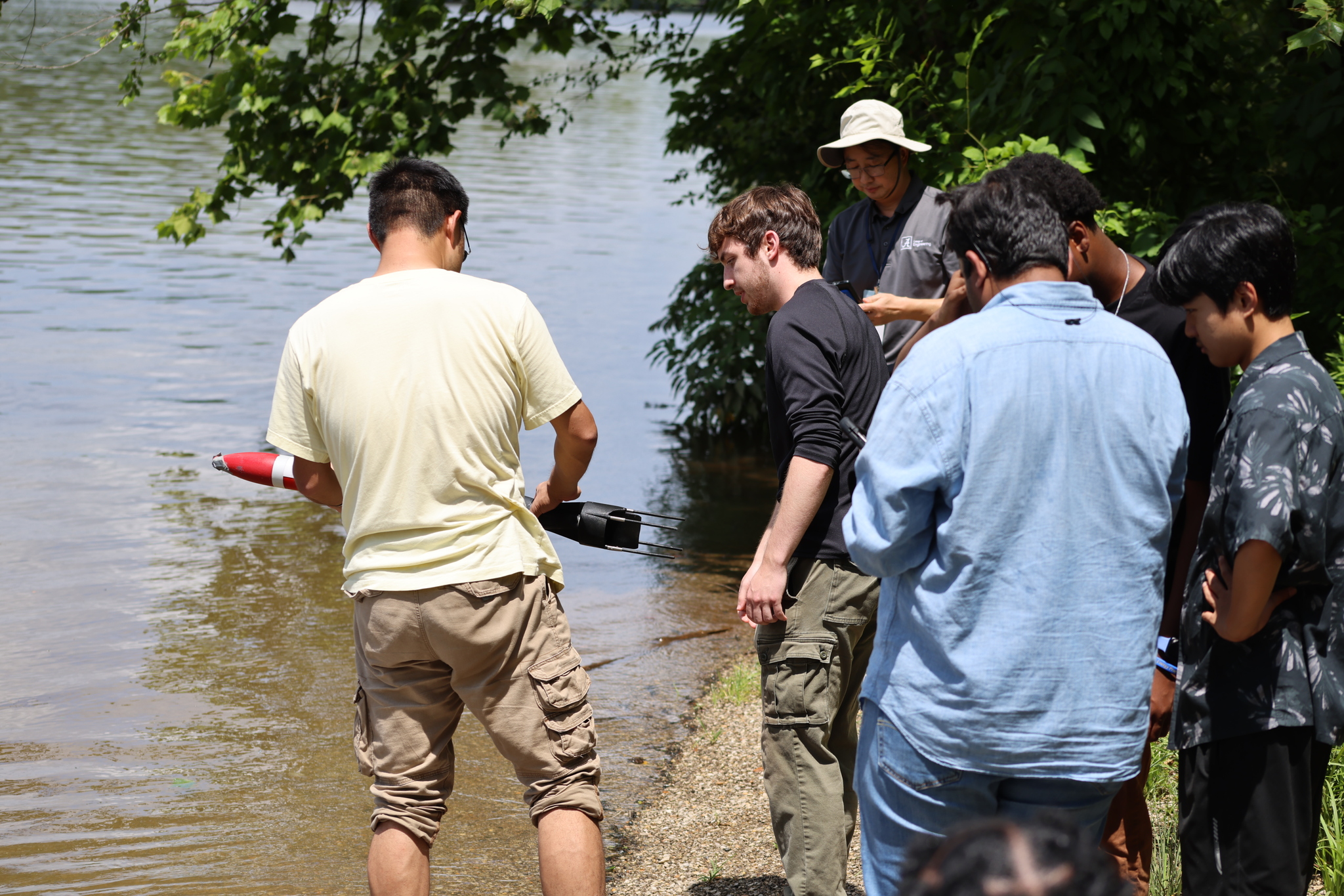 A group of five REU students stand on the edge of the Black Warrior River, observing a research assistant in a light yellow t-shirt demonstrate how the JaiaBot can be deployed.