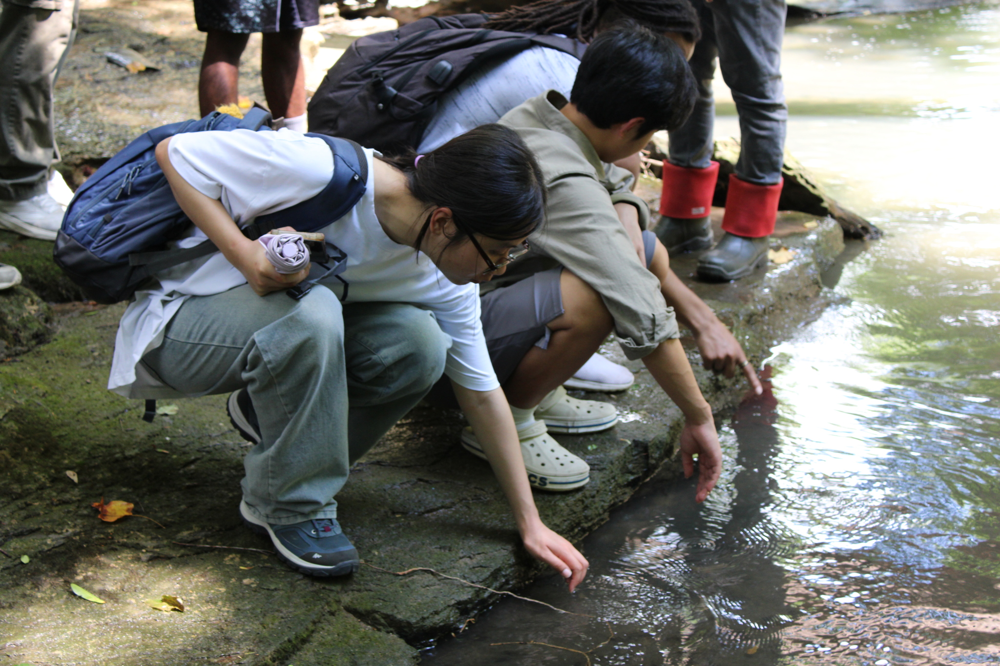 An REU student wearing a white t-shirt kneels by the muddy, rocky edge of Marr’s Spring, reaching her hand into the water. Next to her, another student crouches and points toward the water, while the other students are standing or crouching nearby in the scene.
