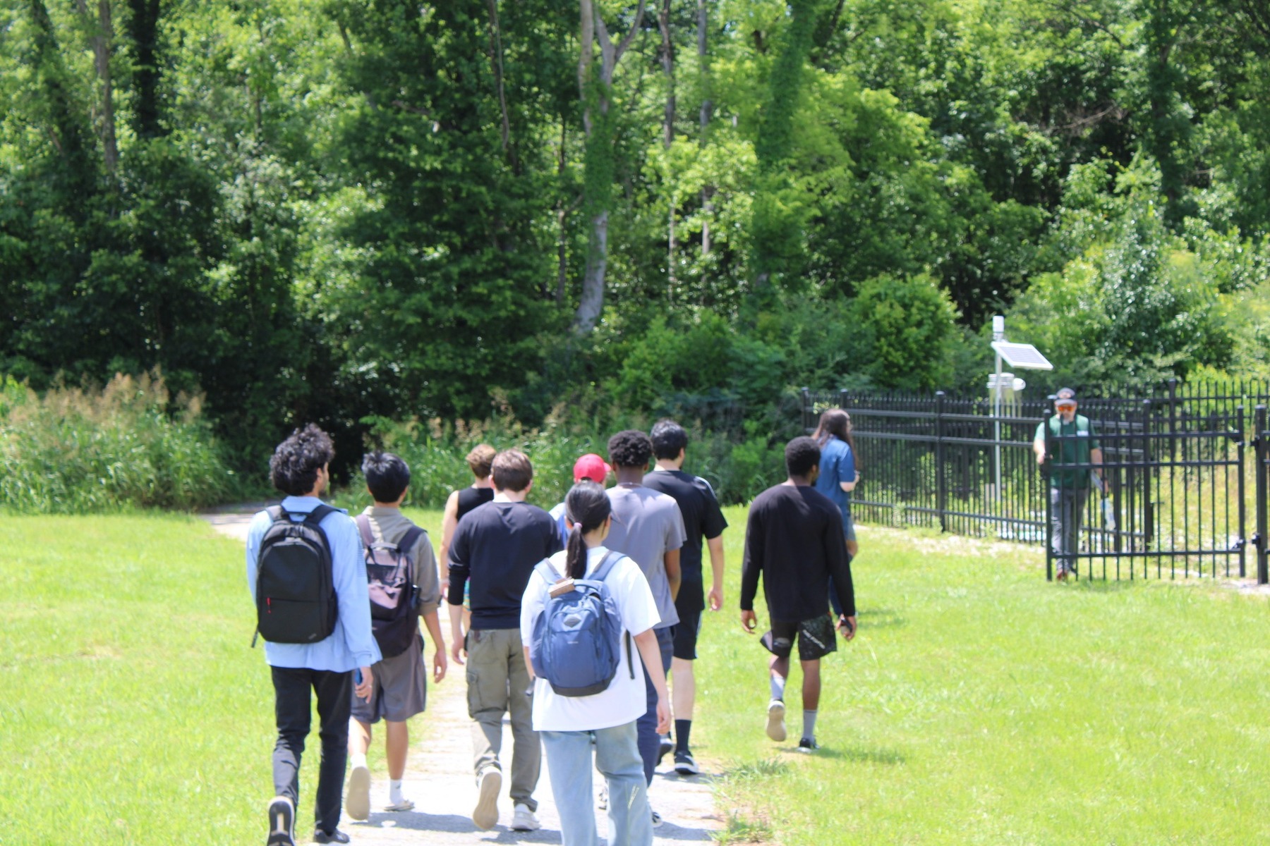 A group of 10 students walk in a line down a grassy trail towards a fenced-in USGS research site where a small solar panel and other equipment are visible.