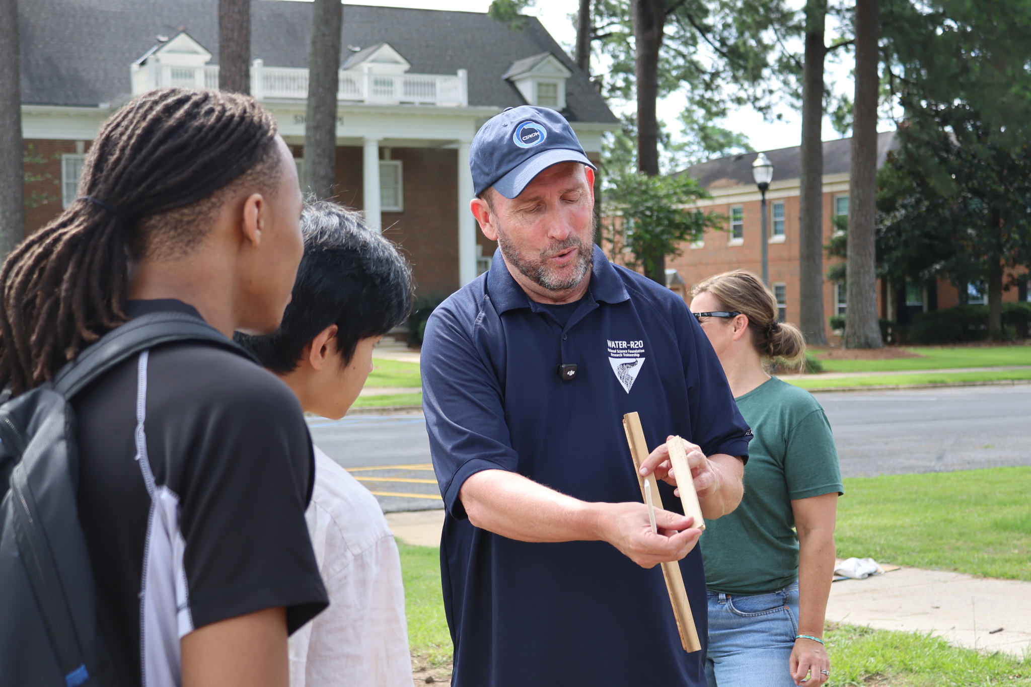 An instructor wearing a blue baseball cap and a dark blue polo shirt holds two pieces of wood, demonstrating the features of the tree rings to two REU students. They are outdoors in the UA campus, with a brick building and large pine trees visible in the background.