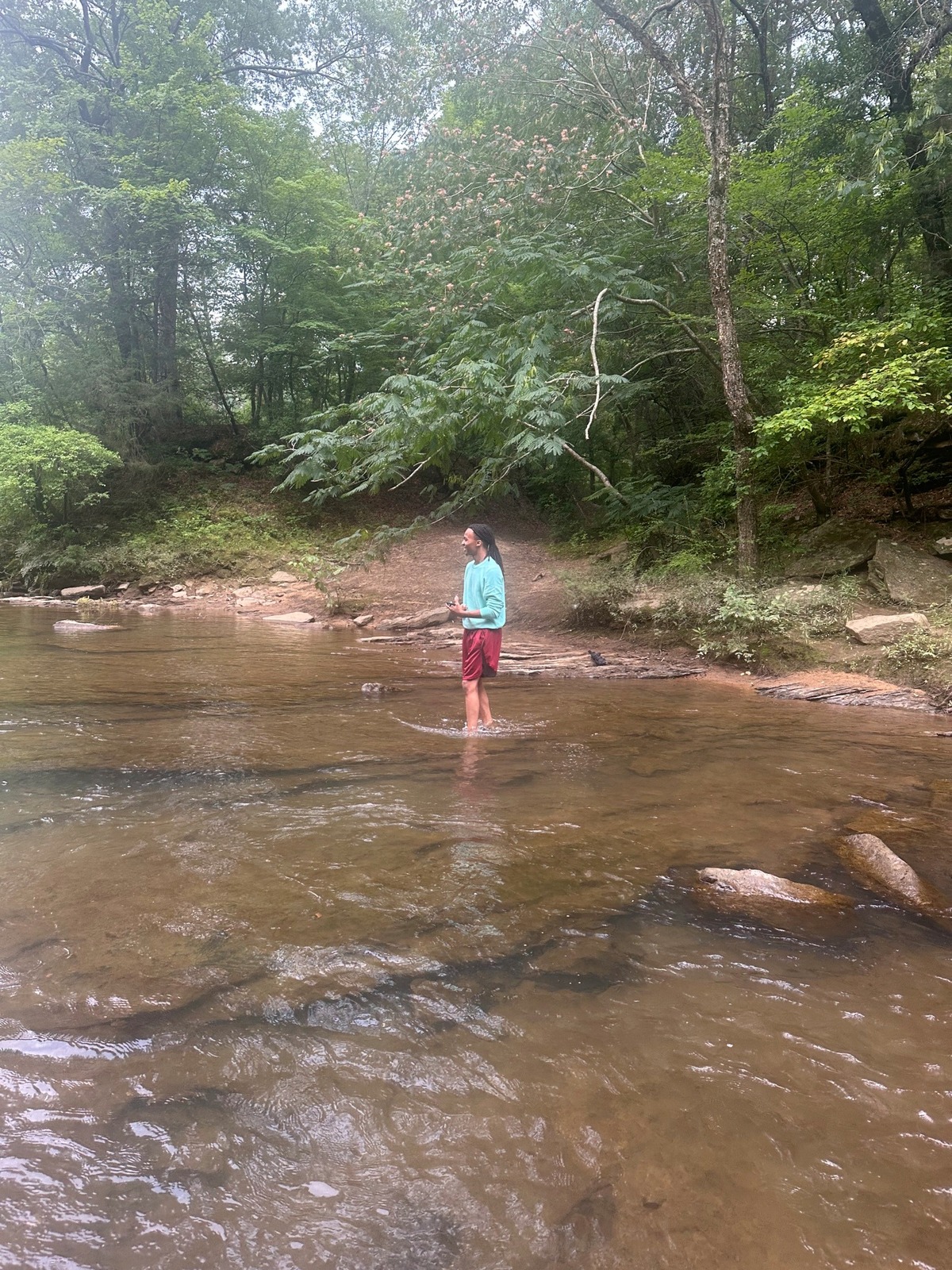 An REU student stands in the middle of a shallow, rocky stream called the Cahaba River, surrounded by a lush forest. He is ankle-deep into the water and smiling away from the camera.