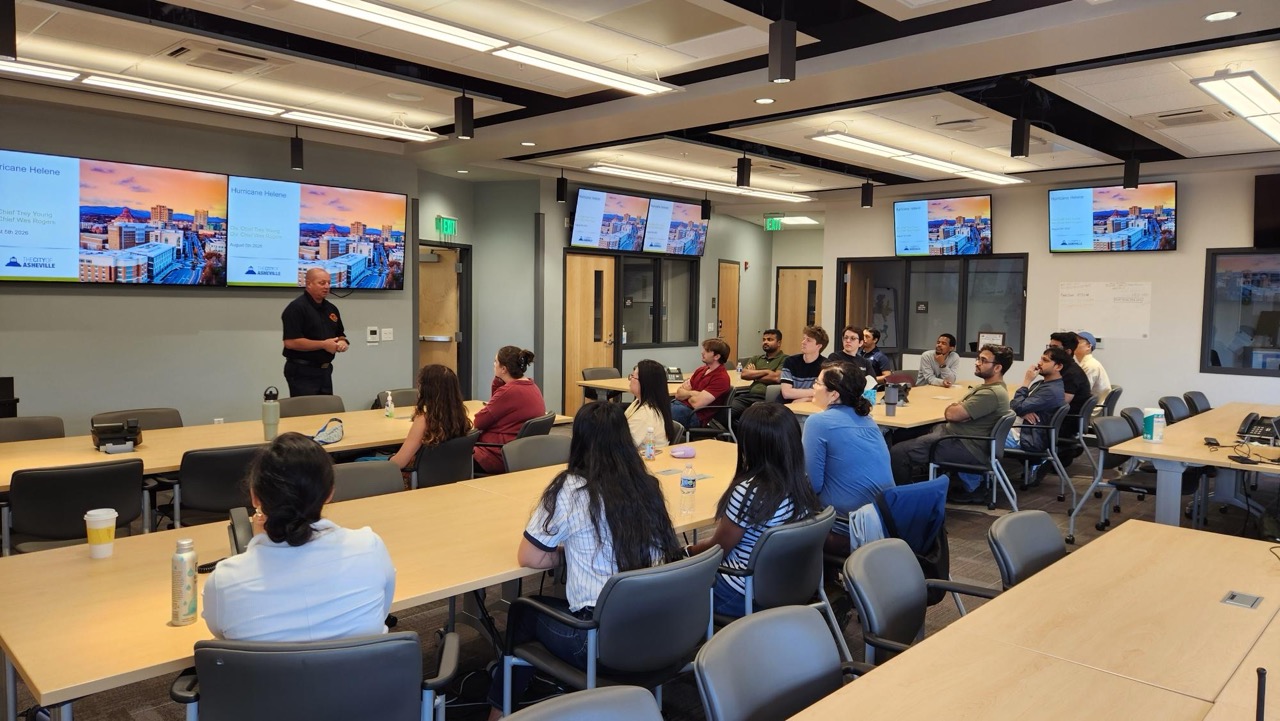 Photograph of a classroom setting with a presenter standing near multiple large screens displaying a presentation slide. Audience members seated at tables face the presenter, with room lighting and modern furniture contributing to a professional learning environment.