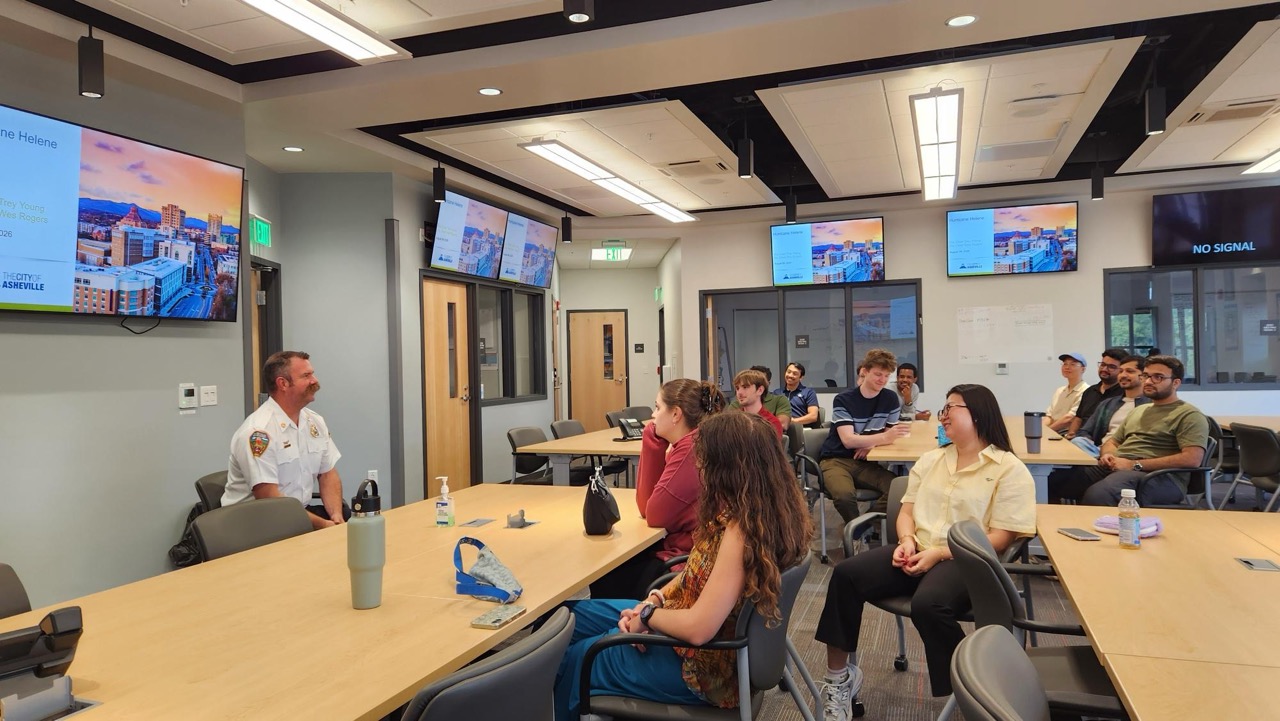 Photograph of a classroom setting showing a group of students seated around tables listening to a speaker in a white uniform at the front. Multiple screens display a presentation slide with a cityscape image and text.