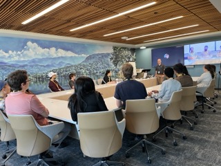 Photograph of a modern conference room with a group of people seated around a large U-shaped table, attentively listening to a presenter standing near a screen displaying a video call with two participants.