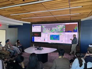 Photograph of a group presentation in a conference room with a large digital screen displaying weather maps, radar data and system status information. Presenter points to colorful maps showing storm patterns and geographic regions, while audience attentively observes around a circular table.