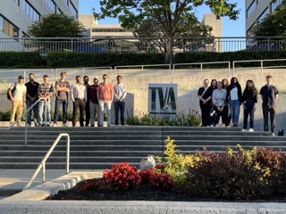 Photograph of a group of 14 people standing in a line on steps outside a building. The scene includes greenery, red flowers in the foreground and modern buildings in the background.