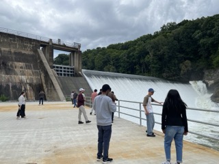 Photograph showing a group of people standing on a concrete platform near a large dam with water flowing over its spillway. Dense green trees surround the dam and cloudy skies are visible above.