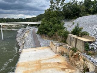 Photograph of a riverside pathway next to a concrete structure with rust stains and rocky embankments. The scene includes a bridge crossing over the river, dense green trees and a cloudy sky.