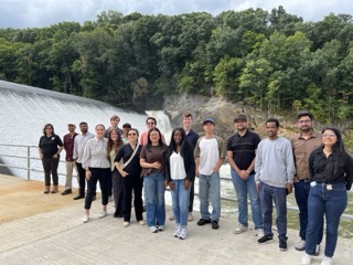 Photograph of a group of 16 people standing on a concrete platform near a dam with water flowing over it and a forested hillside in the background.