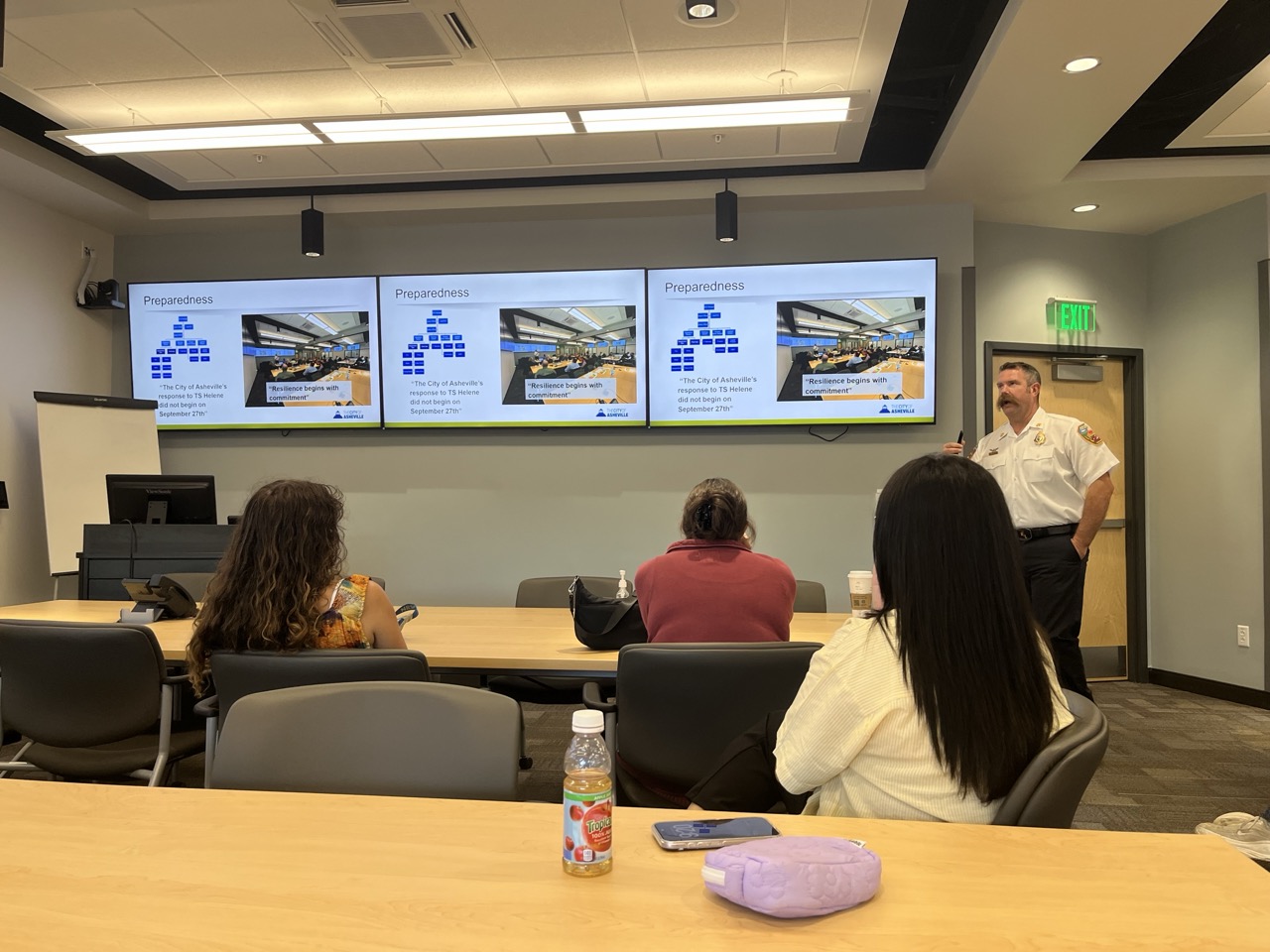 Photograph of a training session in a conference room where a man in a uniform presents preparedness information displayed on three large screens. Screens show a diagram with blue blocks, a photo of a control center and text about city emergency readiness, while attendees sit facing the presenter.
