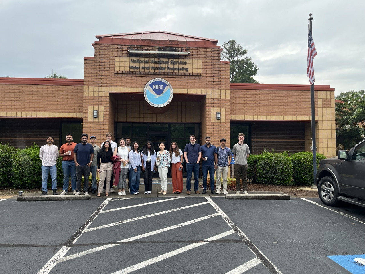 Photograph of a group of people standing in front of a brick building with a NOAA emblem above the entrance. The scene includes a parking lot with marked spaces, an American flag on a pole and overcast sky.