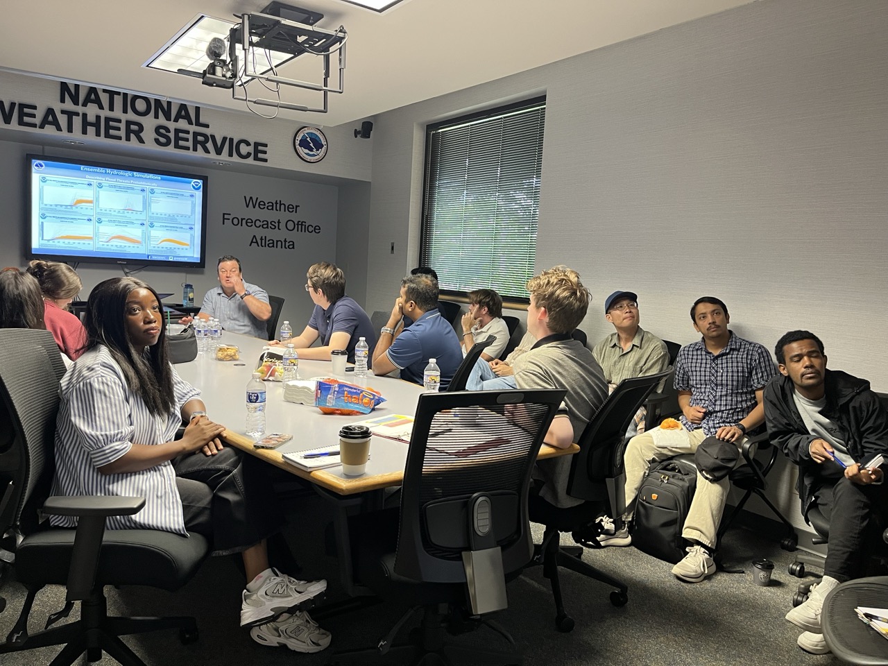 Photograph of a meeting room at National Weather Service Weather Forecast Office Atlanta, showing a group of people seated around a conference table with a large screen displaying weather data and graphs. The room features office chairs, water bottles, snacks and a window with blinds.