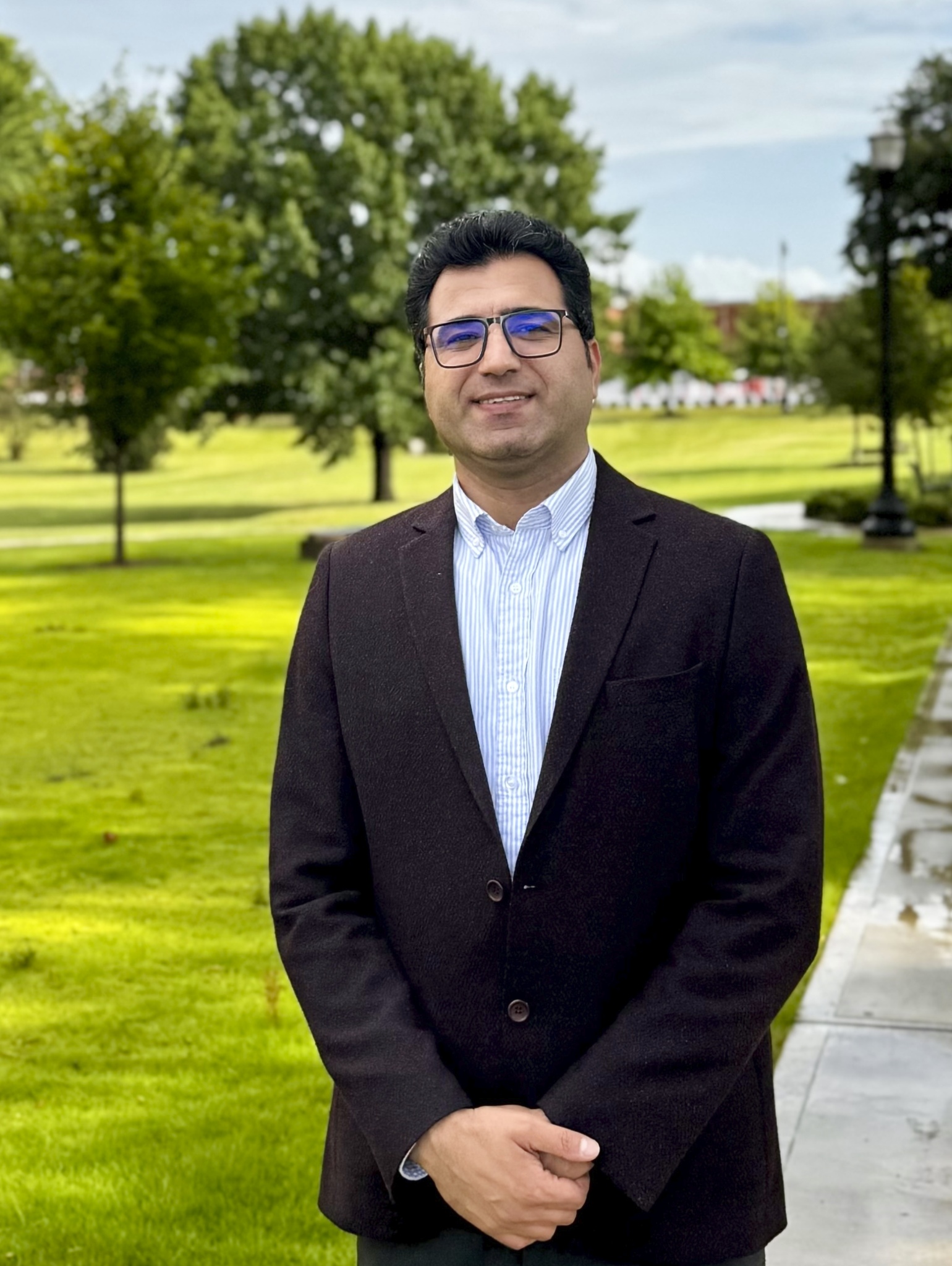 Portrait of Dr. Hazhir Karimi in a black suit and blue-and-white striped shirt, smiling toward the camera and standing outdoors with greenery in the background.